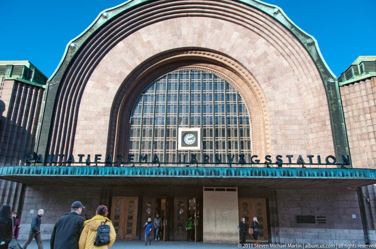 Main entrance of Helsinki Central Railway Station with huge arched window, central clock, bilingual station name, and pedestrians below