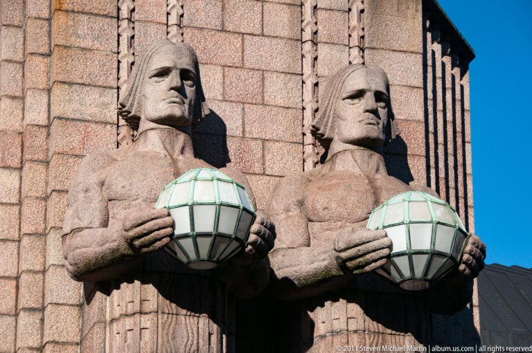 Close view of two monumental granite lamp-bearer figures holding faceted spherical lamps on Helsinki Central Railway Station