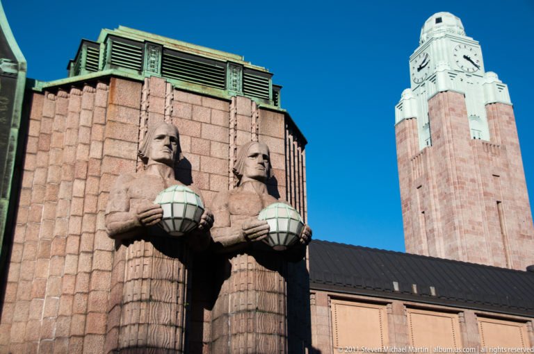 Two granite lamp-bearer statues holding spherical lamps on Helsinki Central Railway Station, with the clock tower rising behind