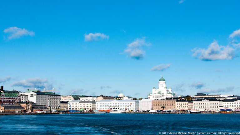 Helsinki Cathedral’s green dome rising above pale waterfront buildings across a broad expanse of blue harbor water.