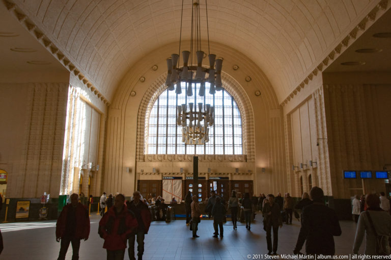 Travelers inside Helsinki Central Railway Station beneath a high vaulted ceiling, chandeliers, and a large arched window