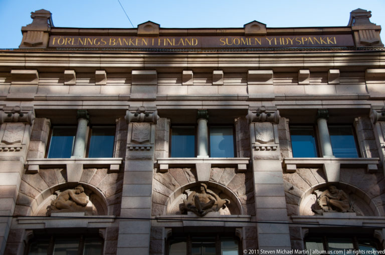 Historic stone bank façade with pilasters, sculptural reliefs, arched panels, and the inscription “Suomen Yhdyspankki” along the roofline
