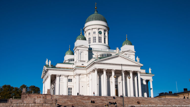 Low-angle view of Helsinki Cathedral showing white neoclassical columns, roofline statues, green domes, and broad stone steps