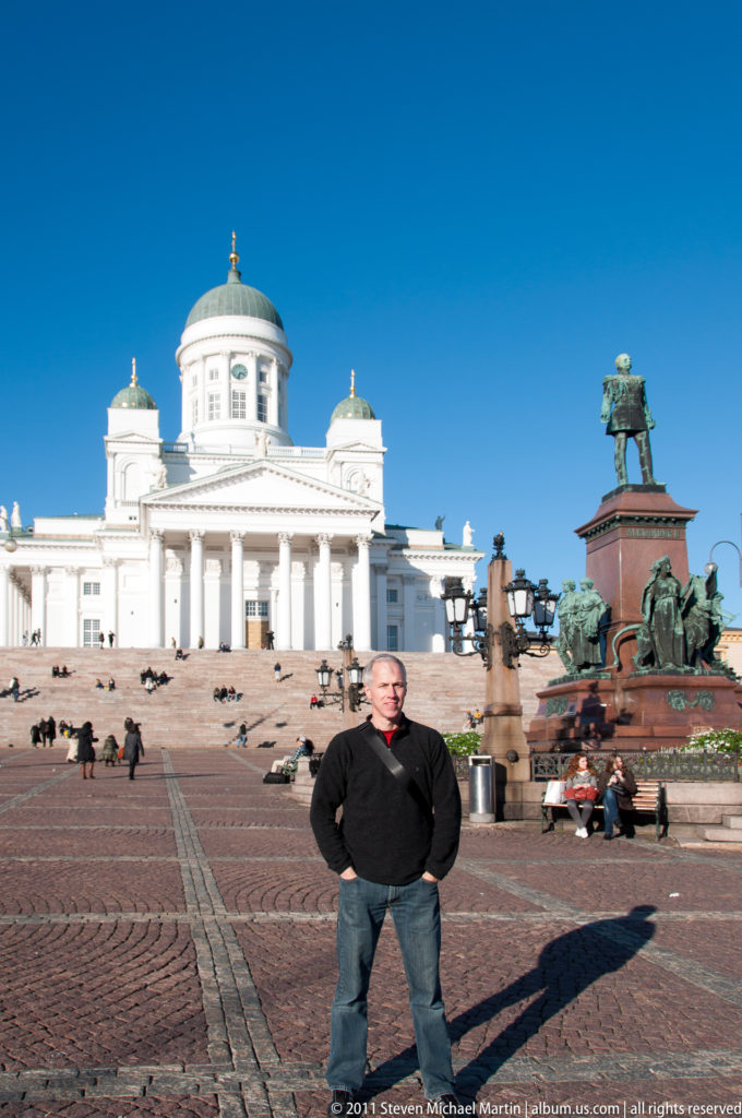 Man standing on cobblestones in Senate Square with Helsinki Cathedral, Alexander II monument, and long shadows behind him