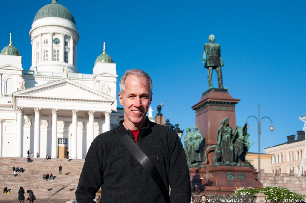 Man in dark jacket standing before Helsinki Cathedral and the Alexander II monument in Senate Square under a blue sky