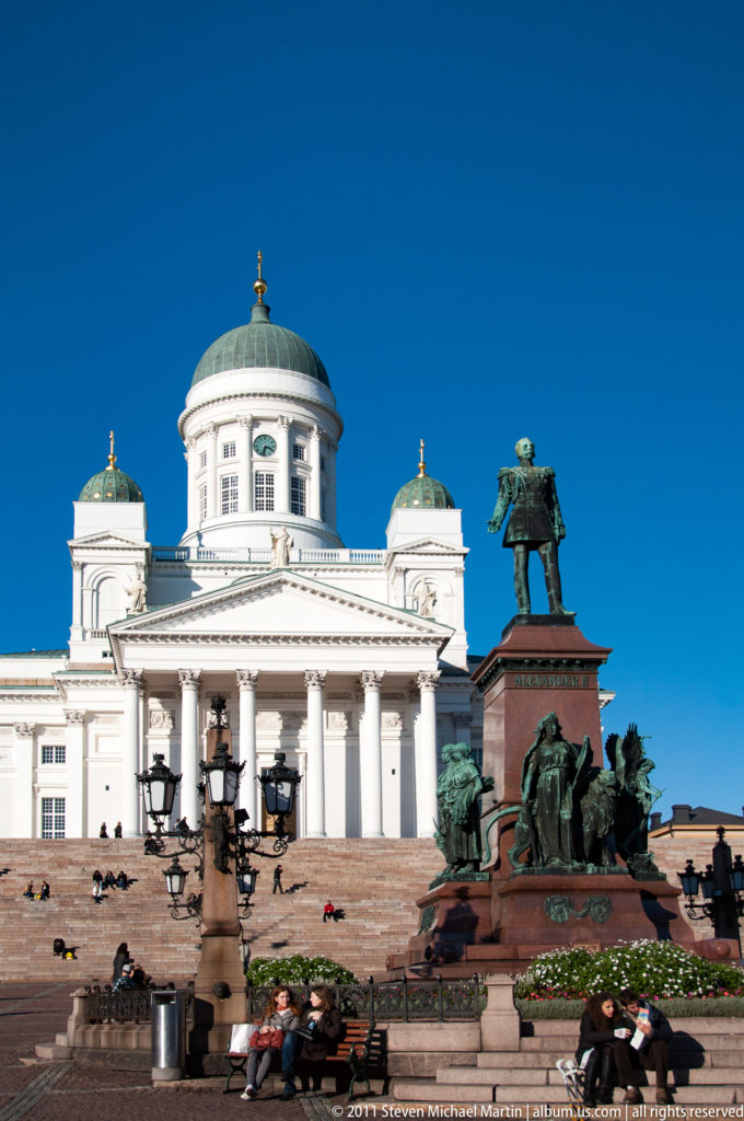 Wide view across Senate Square toward Helsinki Cathedral and the Alexander II monument, with people sitting on steps and benches