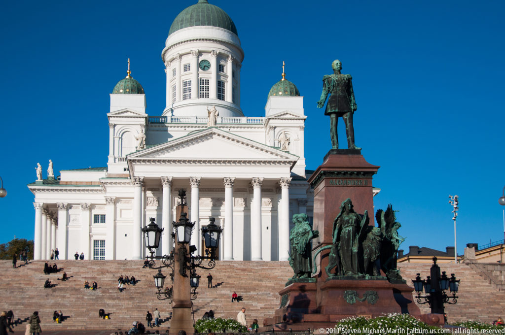 Helsinki Cathedral with white columns and green domes behind the Alexander II monument and visitors on Senate Square steps