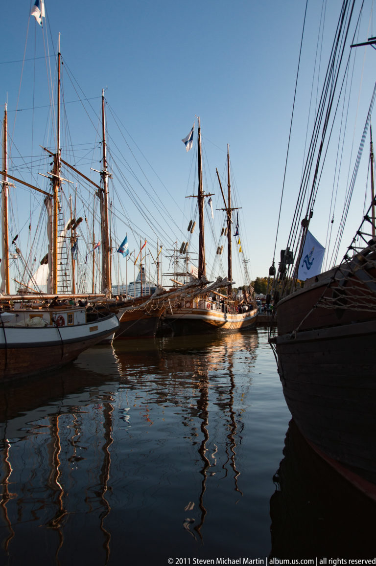 Tall ships and dense rigging line both sides of calm Helsinki harbor water, with dark hulls and reflections in the foreground