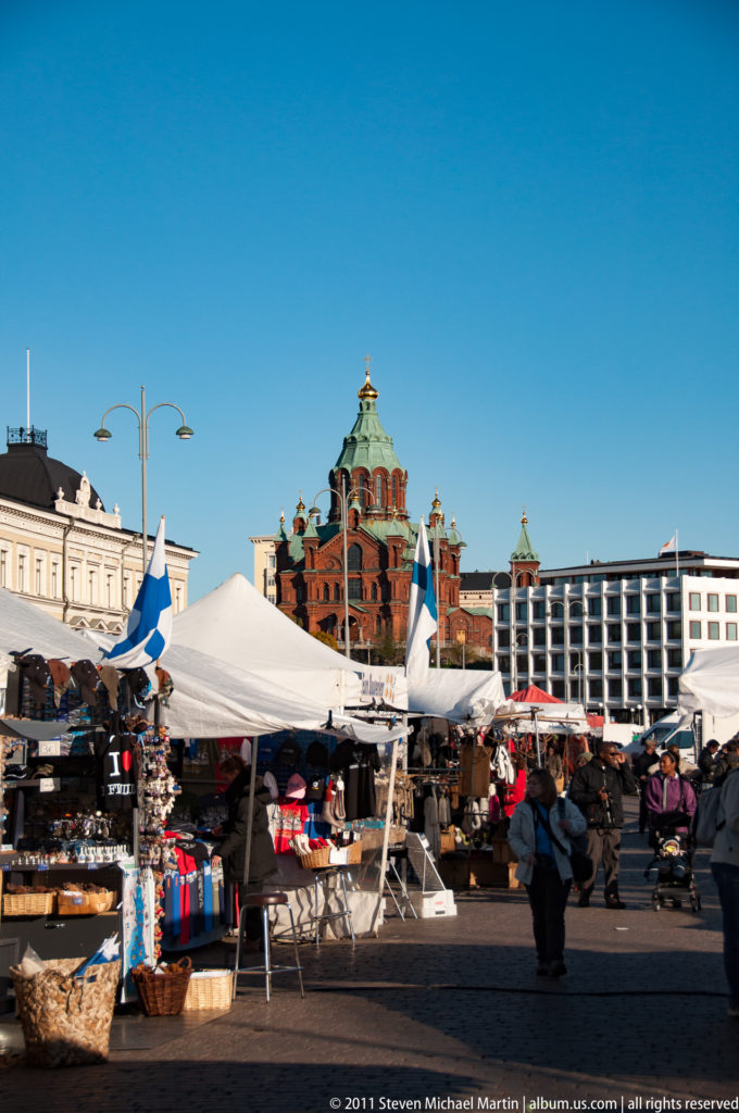 Market stalls, shoppers, and Finnish flags at Helsinki Market Square with red-brick Uspenski Cathedral rising in the background