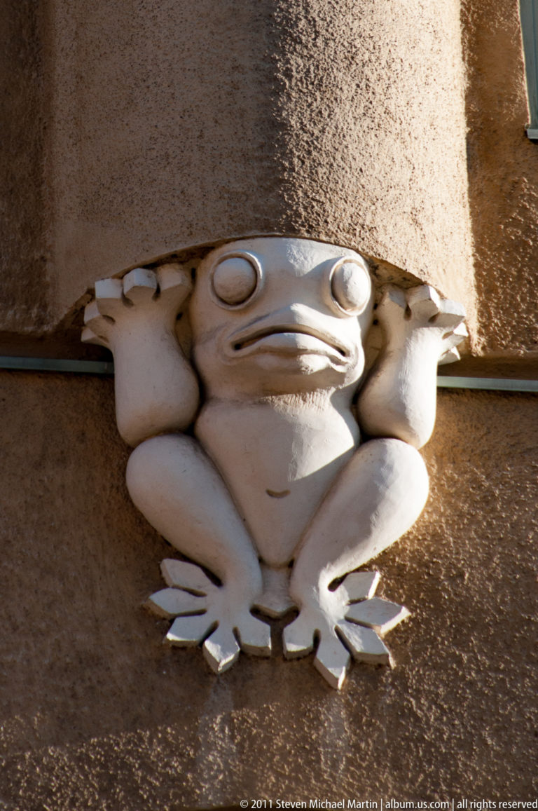 Close view of a pale frog-shaped architectural ornament with round eyes, folded limbs, and splayed feet beneath a stone projection