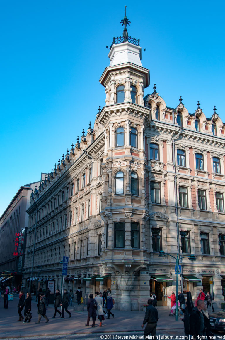 Pedestrians cross a Helsinki street beneath an ornate historic corner building with a rounded bay, turret, dormers, and decorative stonework