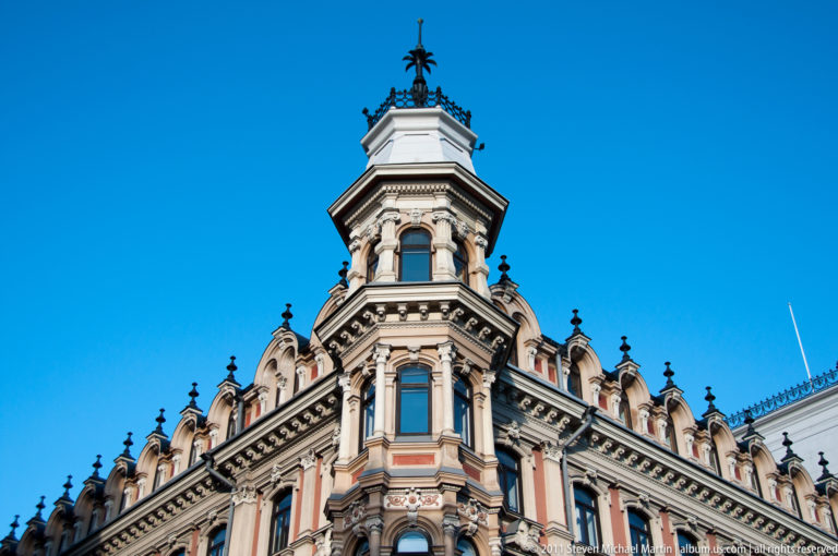 Ornate historic Helsinki building corner with arched windows, columns, dormers, and a tall metal-capped turret against blue sky