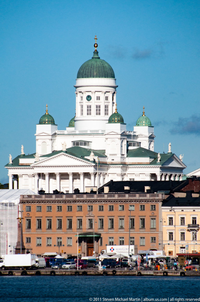 White Helsinki Cathedral with green domes rising above lower waterfront buildings, viewed across the harbor under a clear blue sky.
