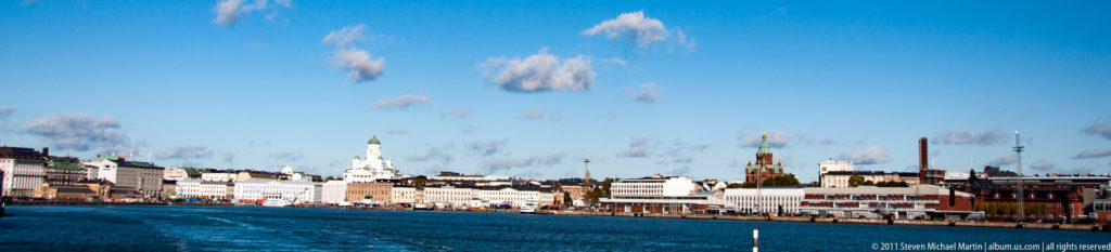 Helsinki waterfront across blue harbor water, with pale city buildings and the green-domed Helsinki Cathedral rising near the center.