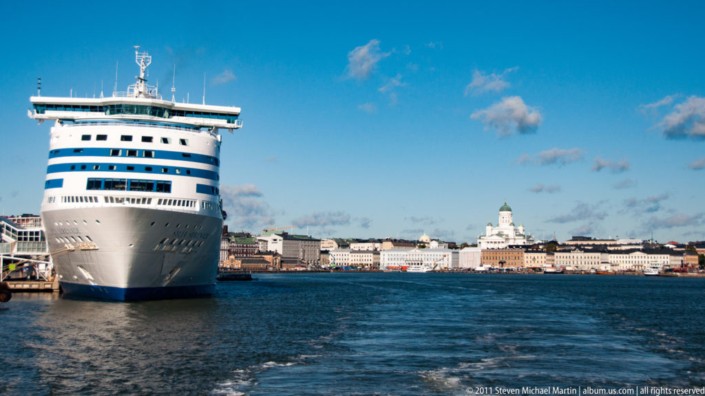 Large passenger ferry seen bow-on in Helsinki harbor, with waterfront buildings and Helsinki Cathedral’s green dome across the water.