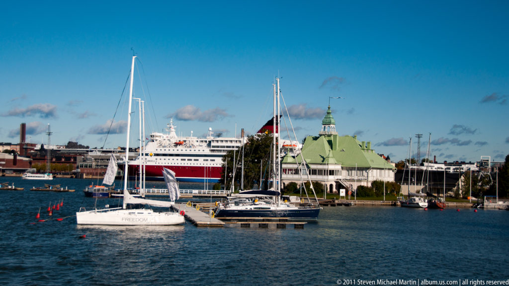 Sailboats moored on blue water before a large red-and-white passenger ferry and a green-roofed waterfront building in Helsinki harbor.