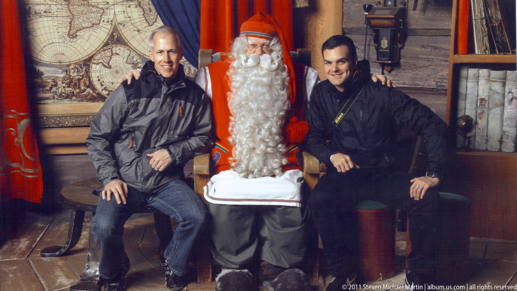 Two men seated beside a costumed Santa Claus performer in a rustic timber interior with red textiles.