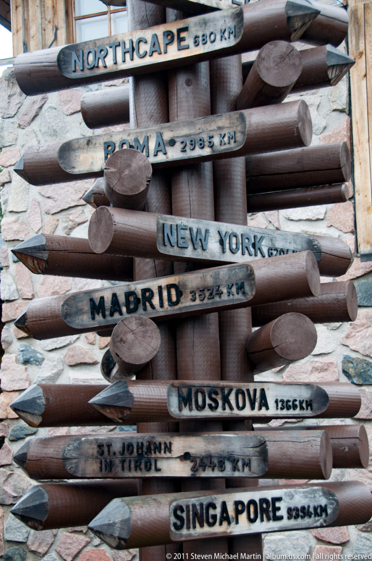 Rustic wooden arrows listing world cities and kilometer distances, including New York, Madrid, Moscow, and Singapore.
