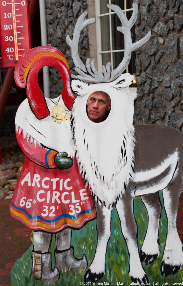 Visitor posing through a painted reindeer cutout beside an Arctic Circle 66° 32′ 35″ sign.