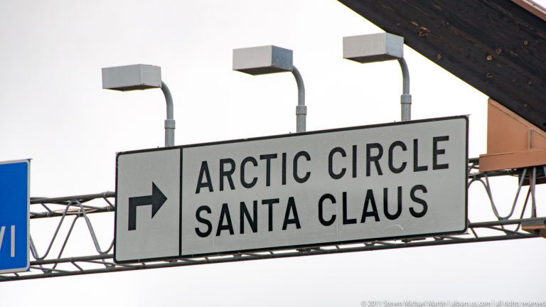 Black-and-white directional sign with a right-pointing arrow and the words Arctic Circle and Santa Claus.