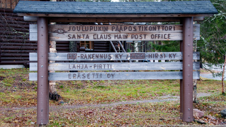 Wooden sign reading Joulupukin Pääpostikonttori and Santa Claus Main Post Office, with additional organization names below.
