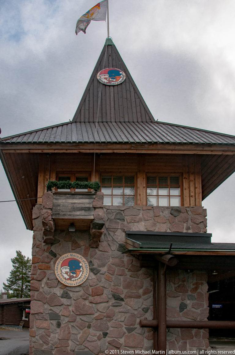 Stone-and-timber façade of Santa Claus Main Post Office in Rovaniemi, with postal emblems and a peaked roof.