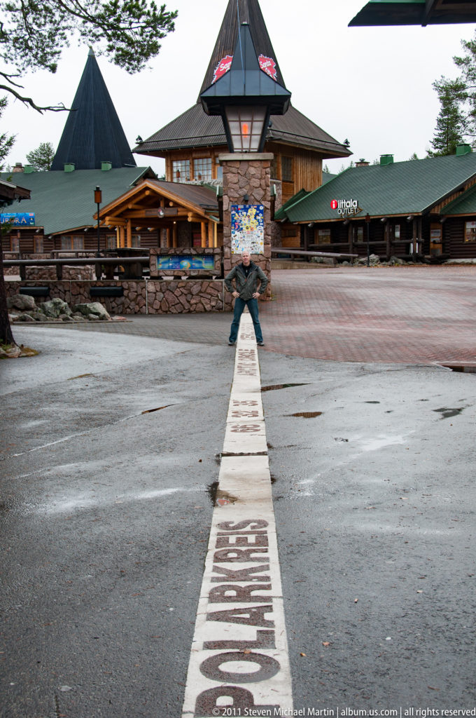 Wide plaza view with a painted Arctic Circle line leading to a stone pillar, timber buildings, and a distant person.