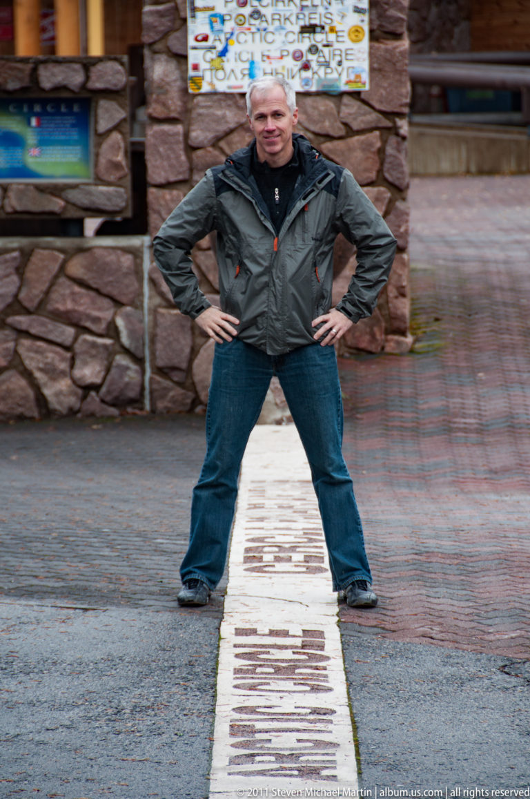 Man standing wide-legged over a painted Arctic Circle line with a stone pillar and buildings behind.
