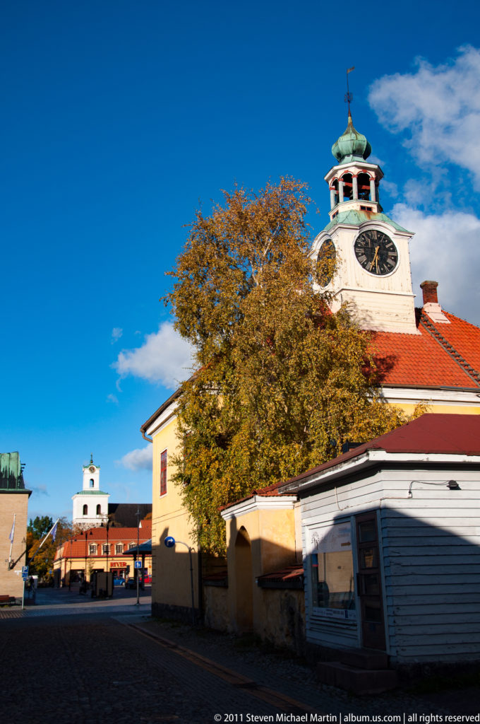 Old Rauma Town Hall clock tower rising behind an autumn tree and low buildings, with blue sky above the market-side roofscape.