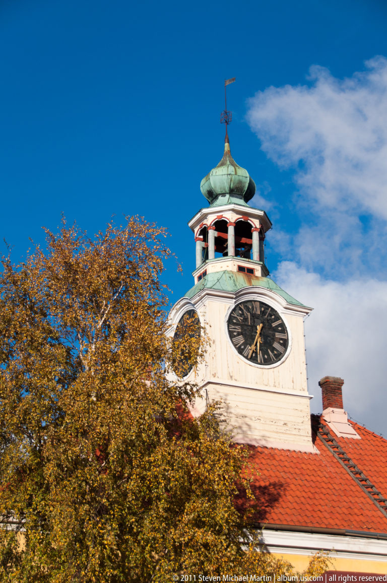White clock tower of Old Rauma Town Hall above red roof and autumn tree canopy, topped by an open lantern and green cap.