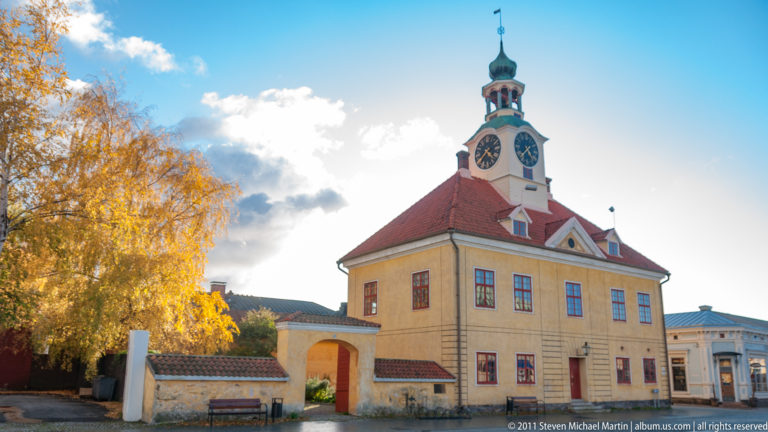 Ochre Old Rauma Town Hall with red tiled roof, white clock tower, green lantern, gateway, and golden autumn birch trees.