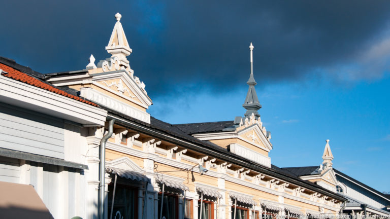 Pale wooden building roofline with decorative gables, tall finials, bracketed eaves, and red and dark metal roof sections under storm clouds.
