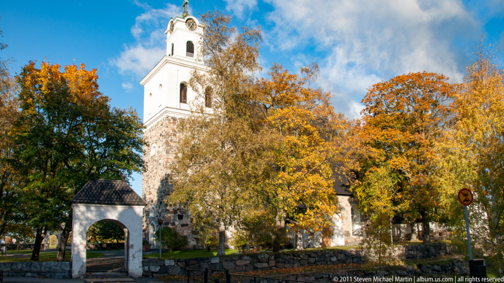 White Church of the Holy Cross tower rising behind orange and yellow autumn trees, with an arched gateway and stone wall in front.
