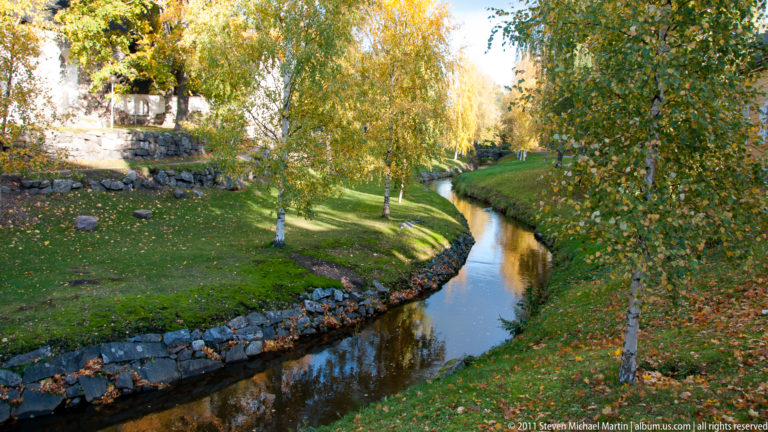 Curving narrow stream bordered by low stones and green grass, with birch trees and golden autumn foliage along both banks.