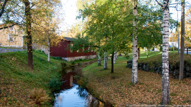 Narrow stream beneath birch trees with fallen autumn leaves, stone edging, grass, and a red wooden building beyond.
