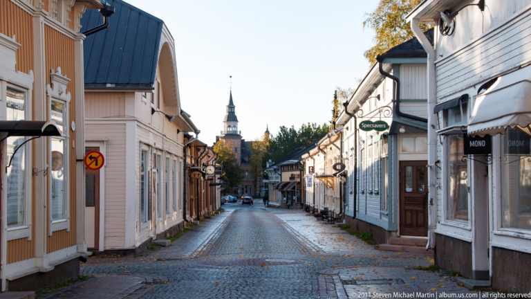 Narrow cobbled street lined with pastel wooden buildings, shop signs, and a church tower in the distance.