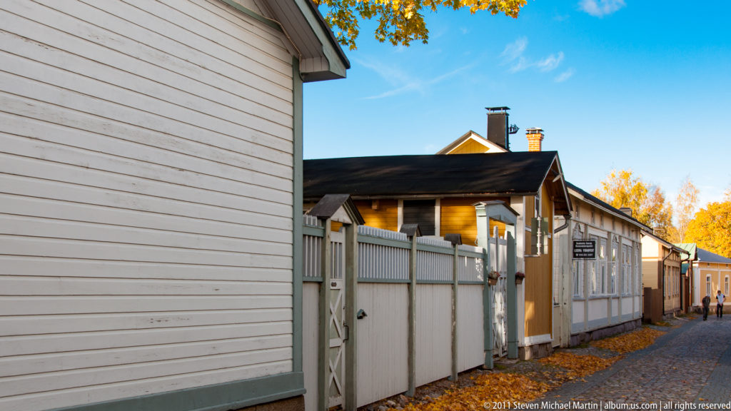 Narrow lane between pale wooden walls and gates, leading past low buildings beneath golden autumn trees in Old Rauma.
