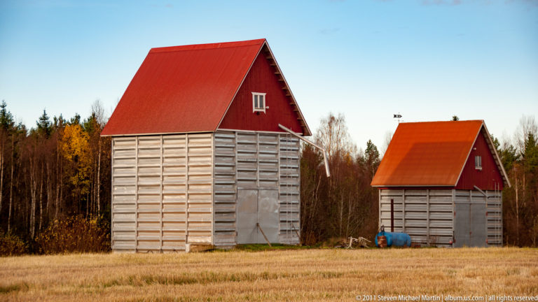 Two pale wooden farm storage buildings with steep red gabled roofs standing in an open field near autumn woodland.