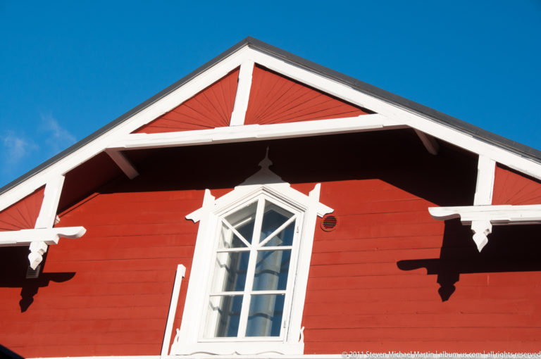 Red wooden gable with a pointed white multipane window, white bargeboards, exposed rafters, and carved brackets against blue sky.