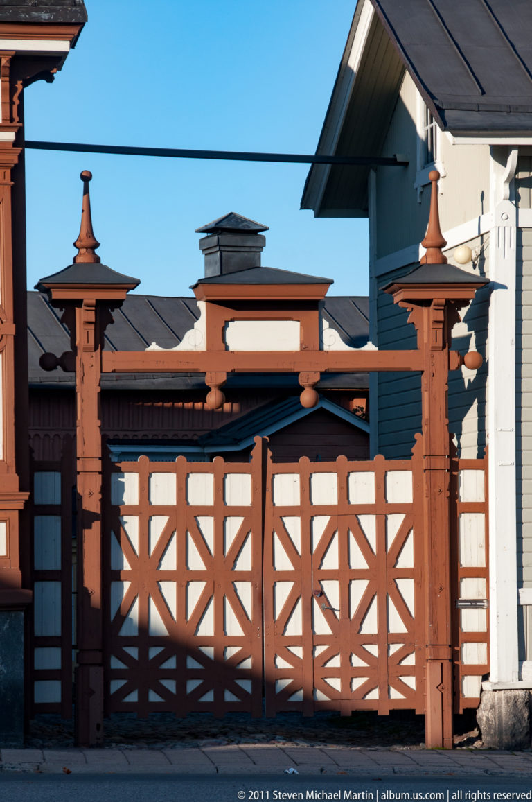 Brown-and-white wooden courtyard gate with diagonal lattice panels, heavy posts, layered rails, and pointed turned finials.