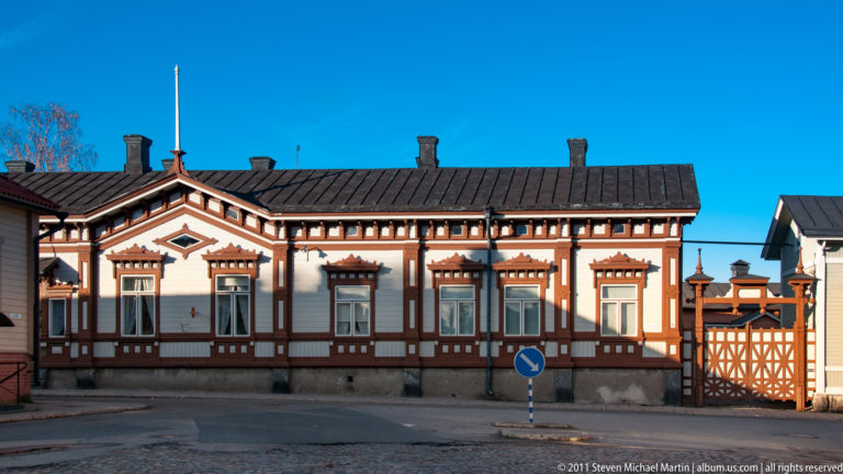 Long white wooden house with brown geometric trim around windows and eaves, plus an ornate matching gate at the right.