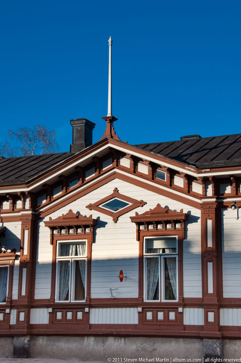 Two tall windows set in a white wooden façade with elaborate brown geometric trim, projecting cornice details, and a slender finial.