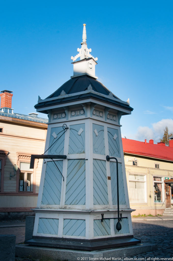 Pale blue polygonal wooden kiosk with closed paneled shutters, dark hipped roof, decorative trim, hinges, and a white finial.