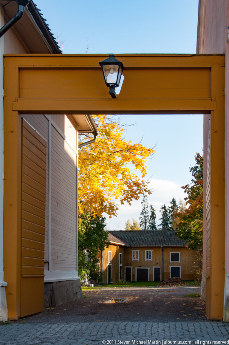 Yellow-painted timber gateway opening onto a cobbled courtyard with a low wooden building and bright autumn tree beyond.