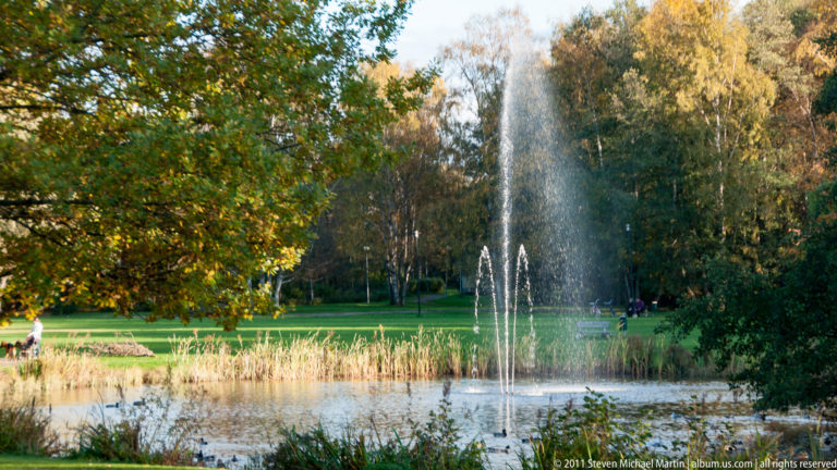 Tall central fountain and smaller water jets in a pond bordered by reeds, lawn, birch trees, and autumn foliage.