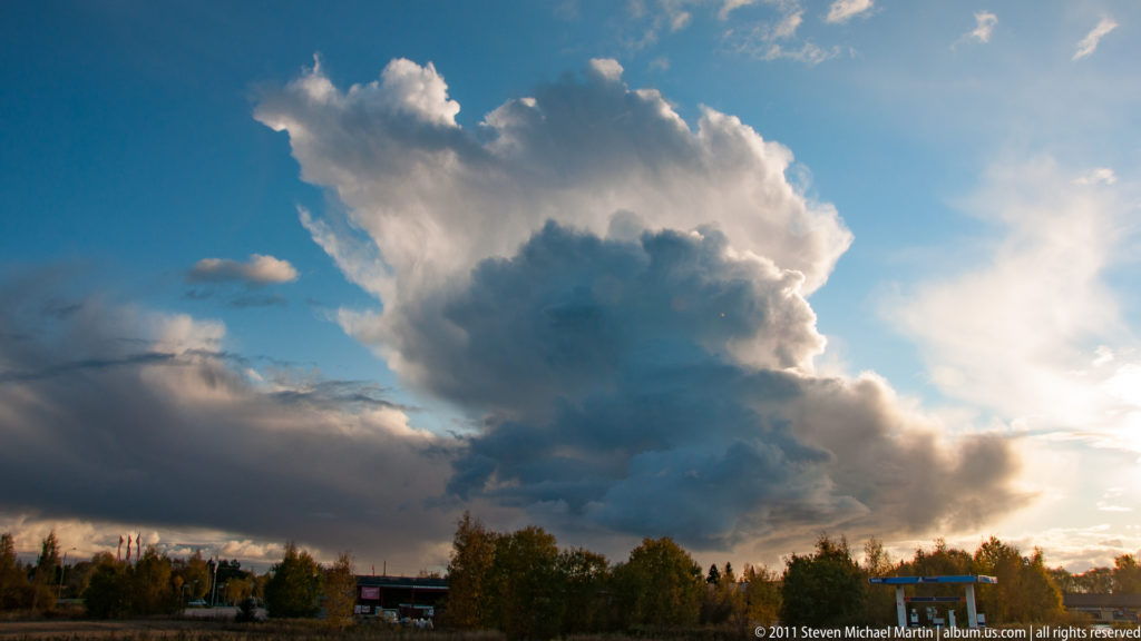 Large sunlit storm cloud rising above a low line of autumn trees and distant buildings beneath a deep blue sky.