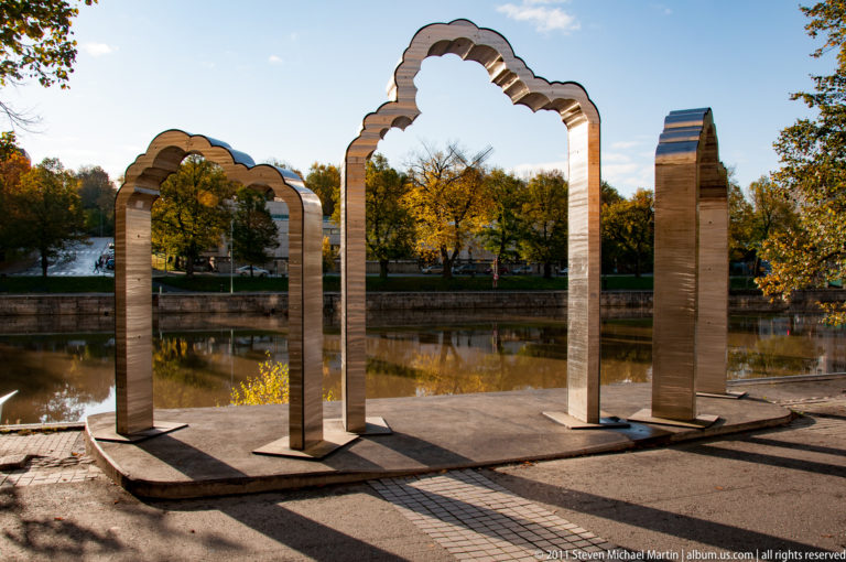 Four freestanding scalloped stone arches on a paved canal terrace, with autumn trees and water visible through the openings.