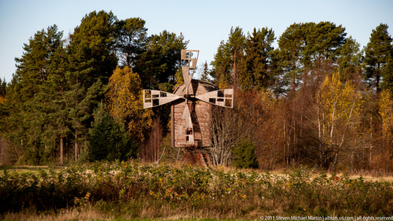 Wooden four-sail windmill standing amid shrubs and meadow with autumn trees and conifers behind it.