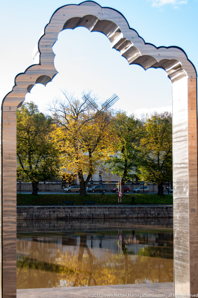 Large scalloped stone arch framing autumn trees, a stone canal wall, and reflections on the opposite bank.