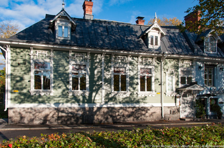 Green wooden house with tall ornate windows, dormers, dark metal roof, and tree shadows across the sunlit façade.